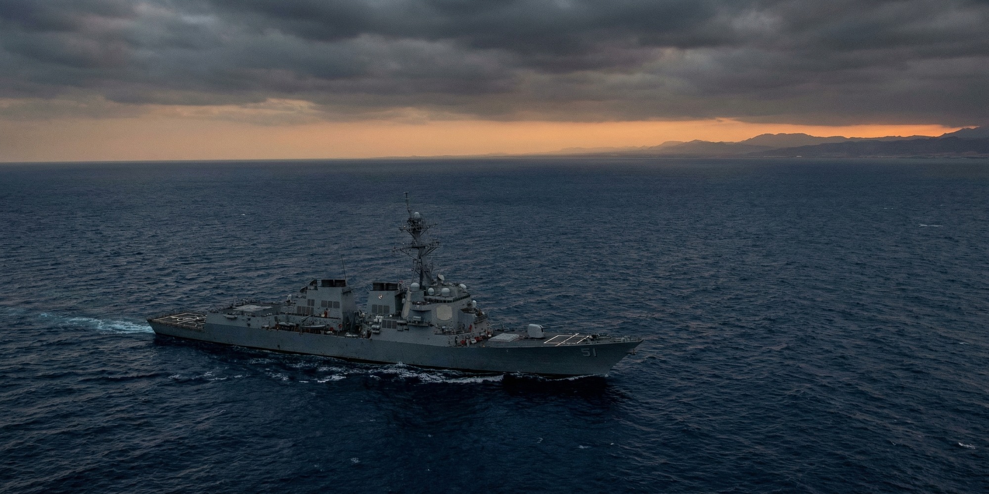 U.S. Navy destroyer underway in the Red Sea at dusk, aerial view, no crew visible, dramatic overcast sky